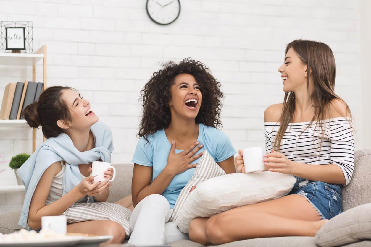 Three women sitting on a couch laughing and drinking coffee.